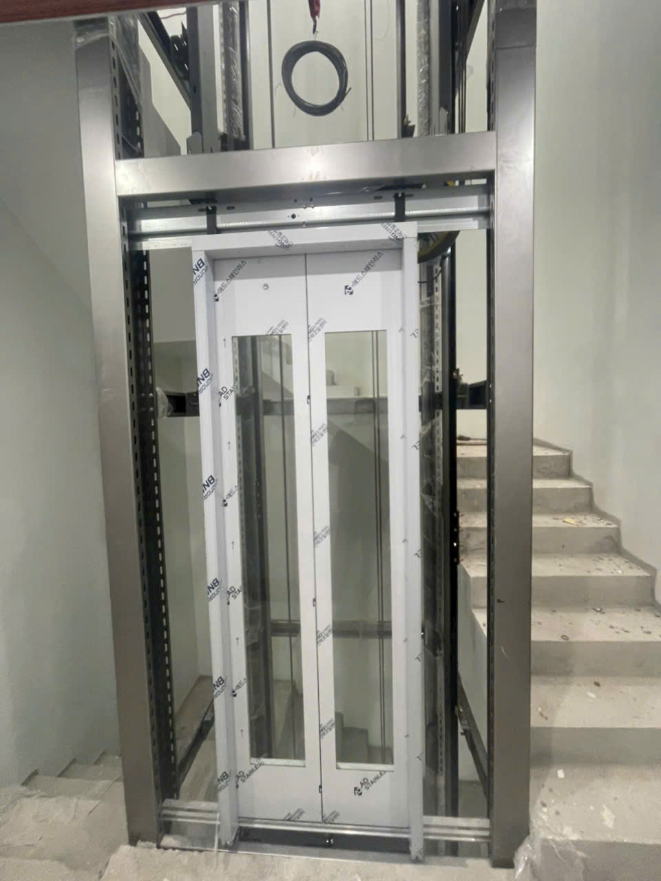 Thang máy gia đình khung kính đang lắp đặt giữa hai nhánh cầu thang trong nhà